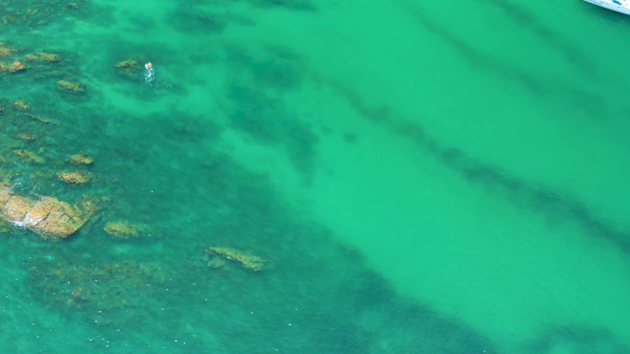 Top-down Palmilla Beach water view with dark reef forms below the surface, tan rocks at left, and a partial white boat near the upper right edge, no visible people.