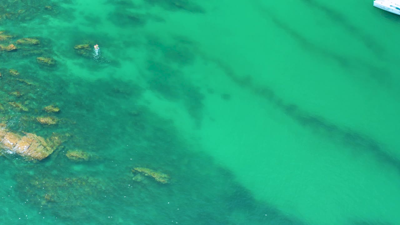 Overhead Palmilla Beach water with reef and rocks along the left edge, open green water across the frame, and a small sliver of white boat at the top, no visible people.