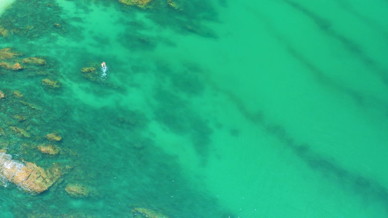 Opening overhead frame at Palmilla Beach showing turquoise water, submerged rocks near the left edge, darker underwater patches, surface ripples, and no clearly visible people.