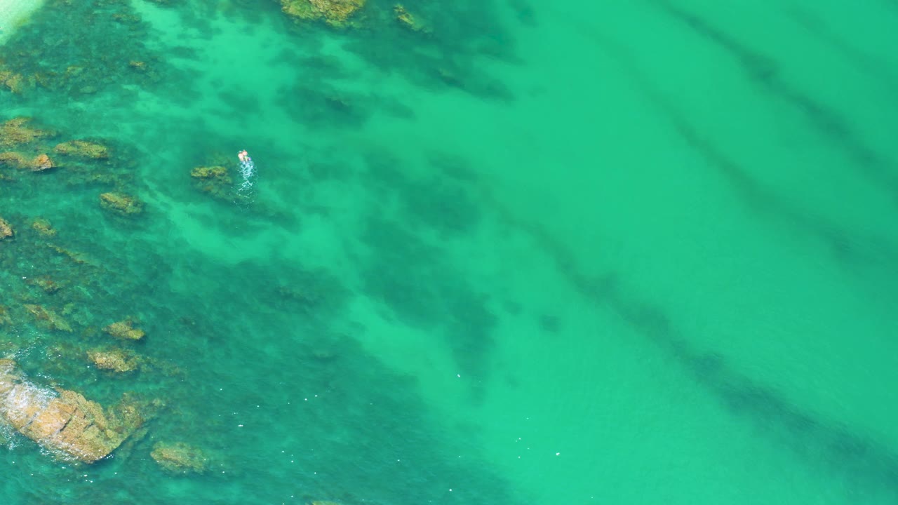 Palmilla Beach aerial frame showing transparent green water, scattered submerged rocks in the upper and left portions, darker reef-like shapes, and no clearly visible people.