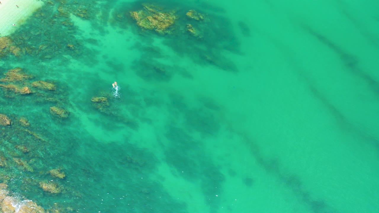 Aerial frame over Palmilla Beach turquoise water with submerged rocks at the left and upper edge, darker reef-like patches below the surface, and no visible people.