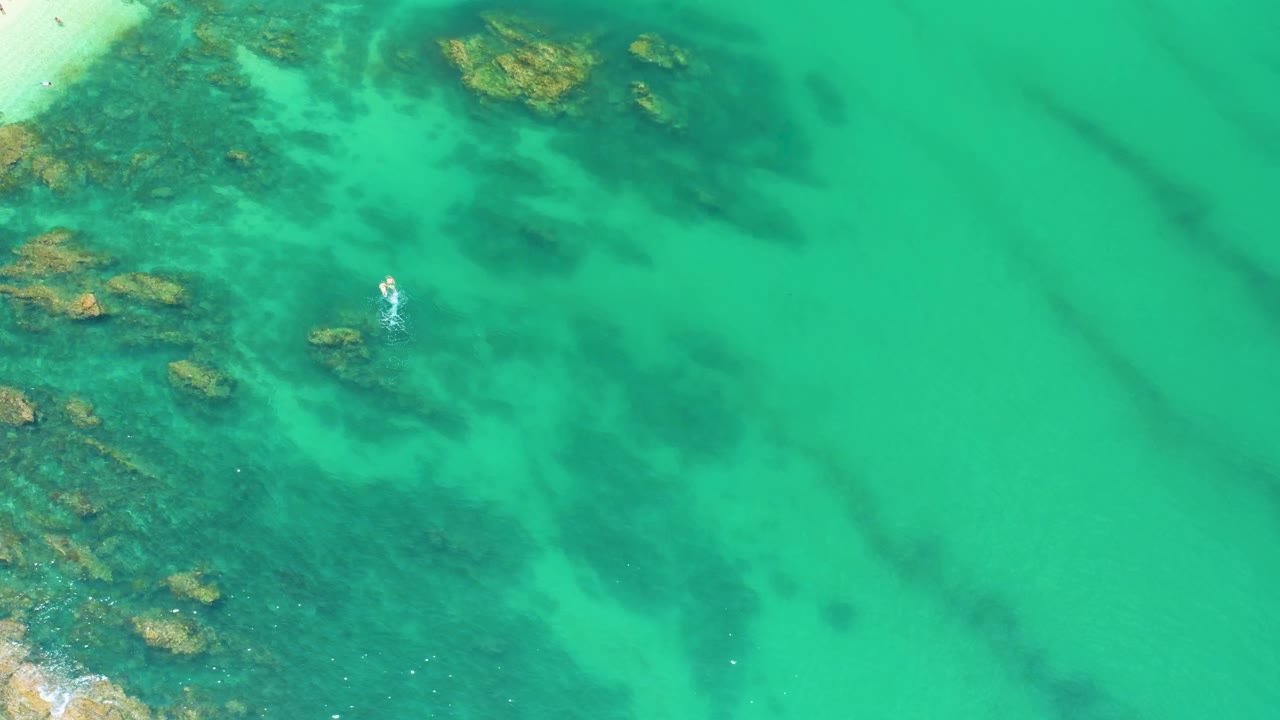 Overhead view at Palmilla Beach showing green sea, clustered submerged rocks on the left, a small white surface disturbance near center-left, and no visible people.
