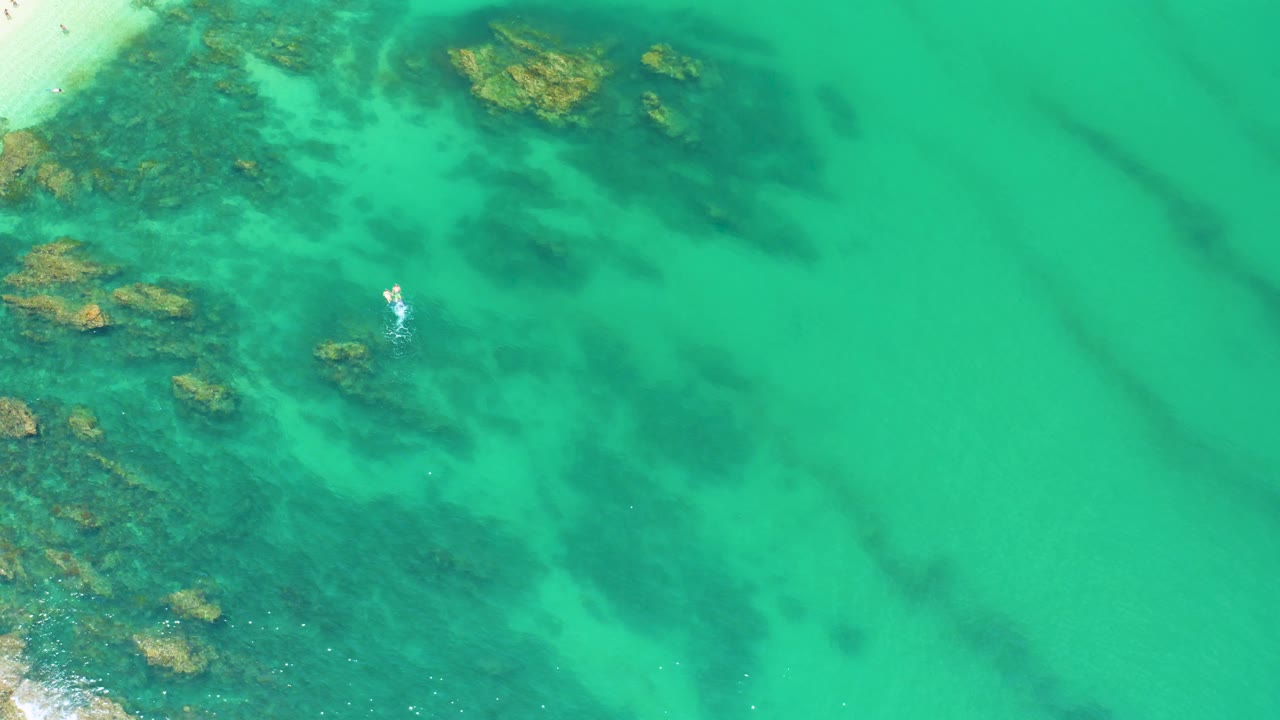 Aerial Palmilla Beach frame with clear green water filling most of the image, rocky shallows along the left and top, faint dark underwater streaks to the right, and no visible people.