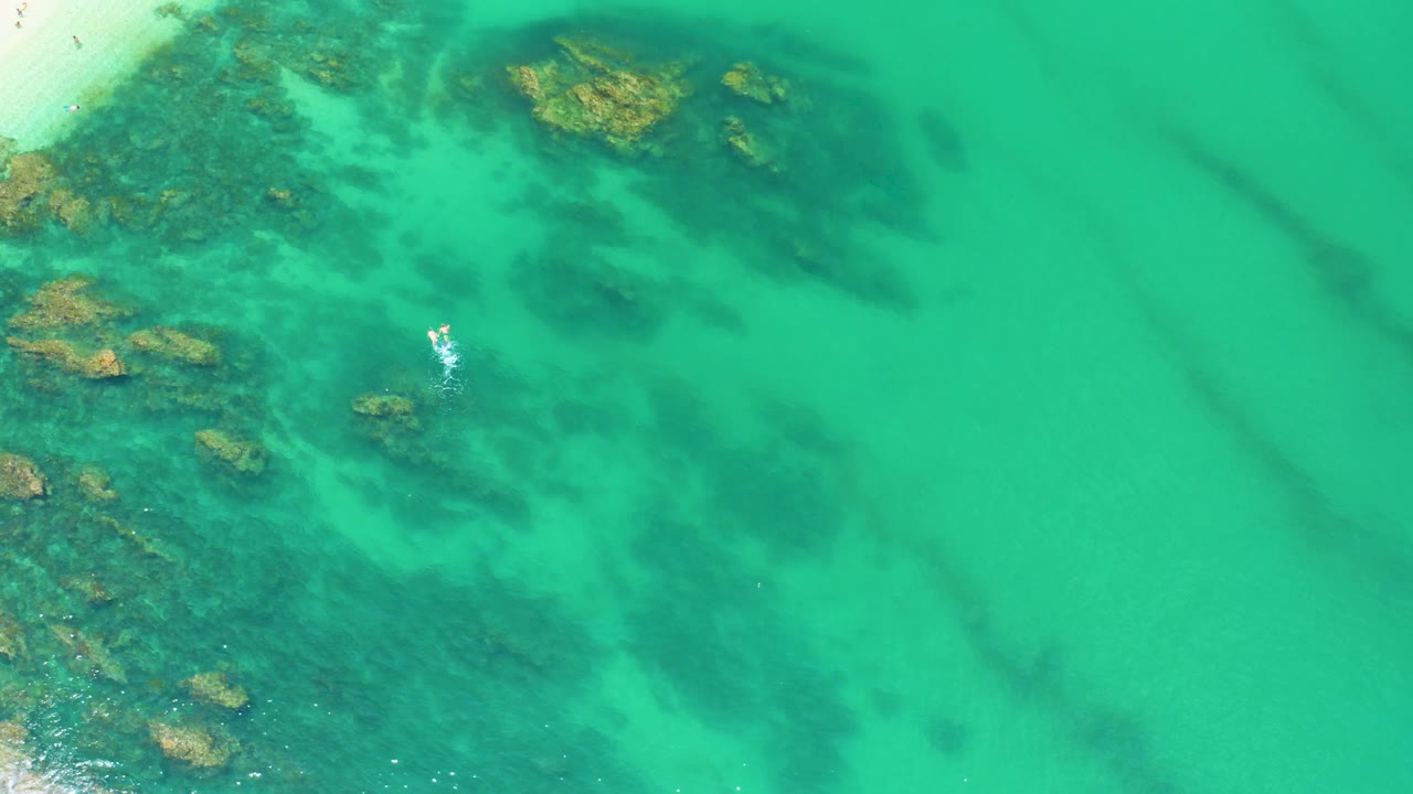 Palmilla Beach aerial frame with reef-like dark patches spread under turquoise water, exposed-looking rocks at the left edge, a small white surface mark, and no visible people.