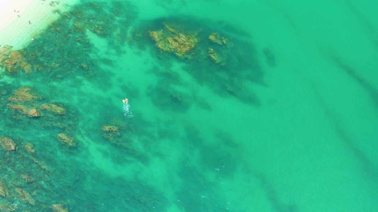Overhead view of blue-green water at Palmilla Beach, Baja California Sur, with dark submerged rock patches, a pale sand edge at upper left, and no visible people.