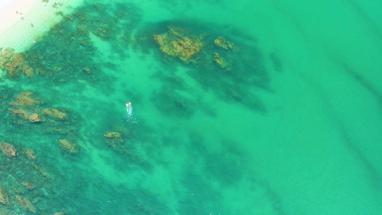 A top-down view over Palmilla Beach water shows clustered dark submerged rocks on the left and upper center, open turquoise water on the right, and no visible people.