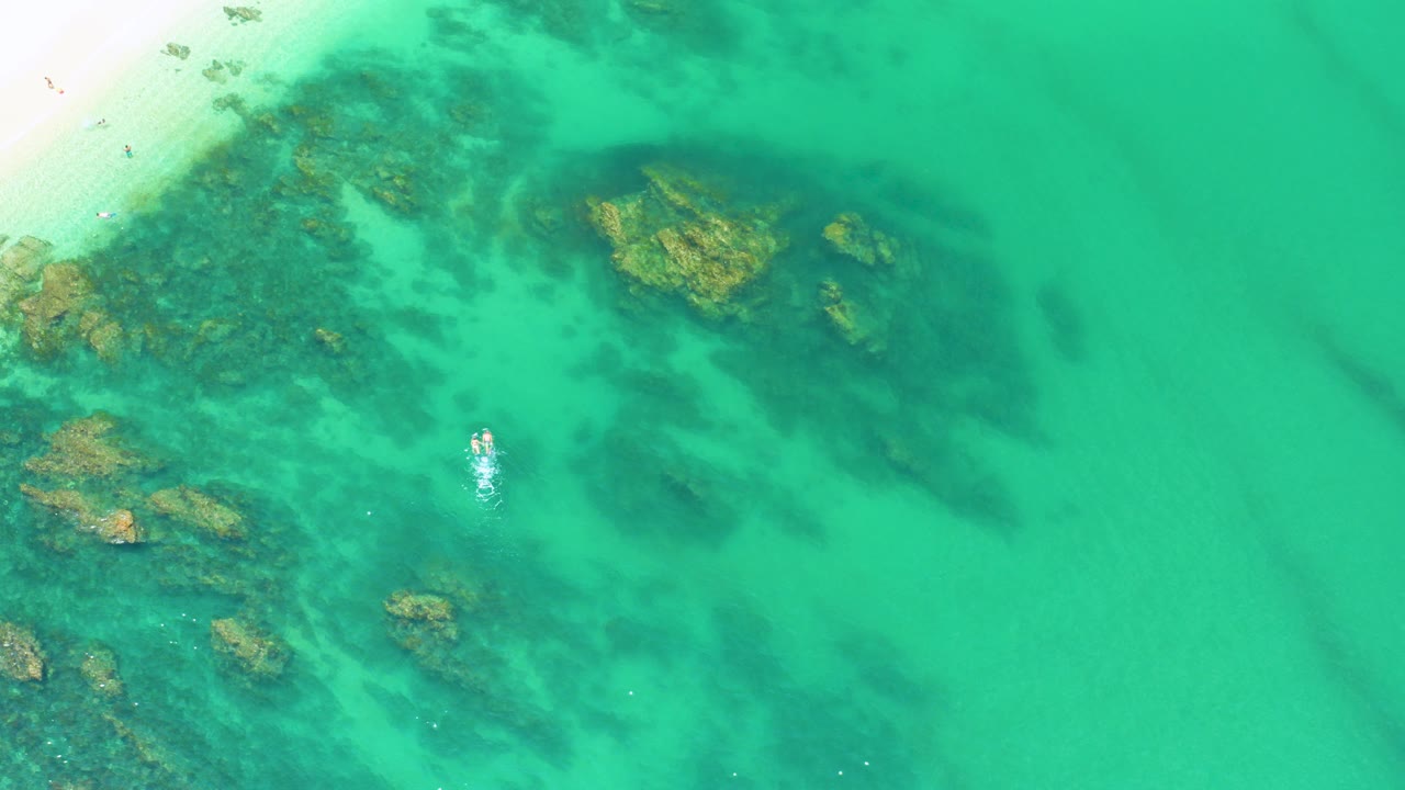 Overhead view of Palmilla Beach water with dark algae patches, tan rocks, pale sand at the upper-left edge, small white wave marks, and no visible people.
