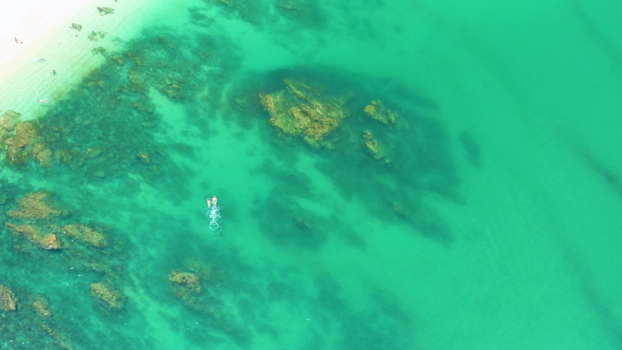 Top-down Palmilla Beach frame showing bright turquoise water, rock formations near the beach side, dark algae shapes beneath the surface, and no visible people.