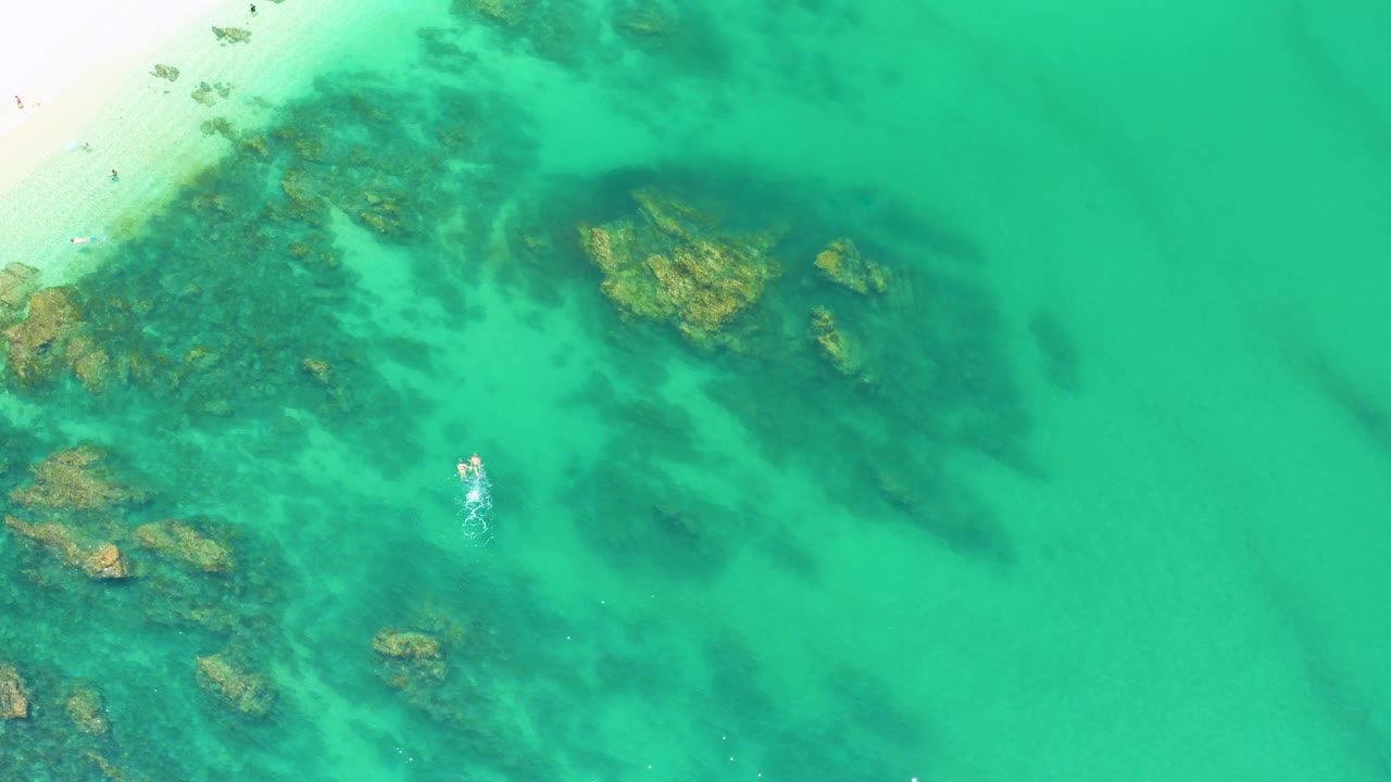 Turquoise ocean surface with dark algae fields and rocks along the left side.