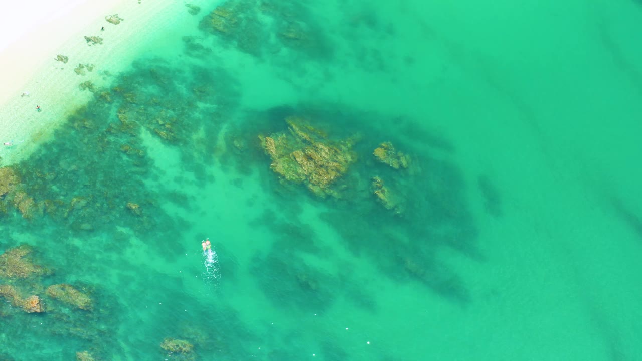 Clear green-blue sea over dark rocks with beach at upper left.