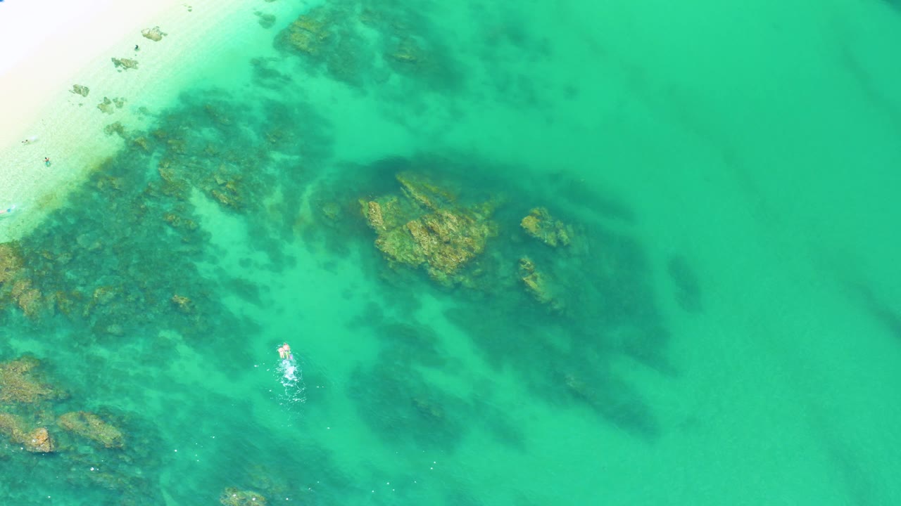 High overhead frame of Palmilla Beach with pale sand in the upper left corner, translucent turquoise water, dark reef-like rock patches, and a small distant person near the shoreline.
