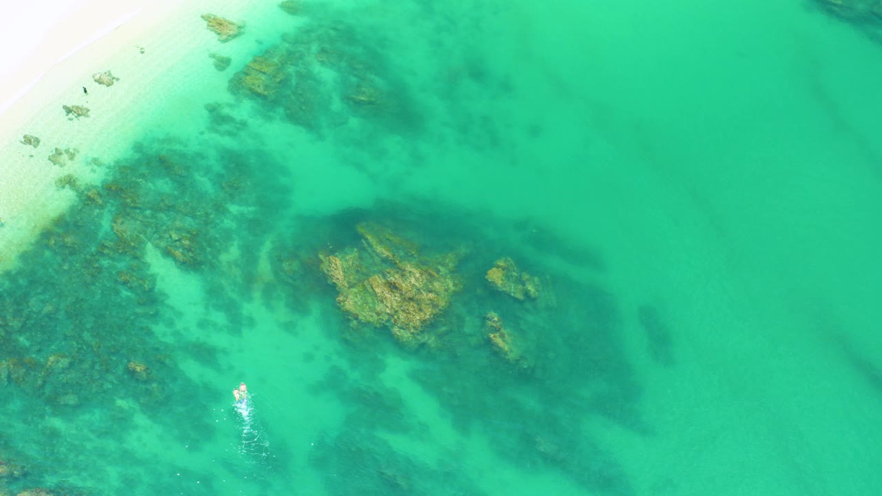 Overhead turquoise water with submerged rocks and pale beach sand.