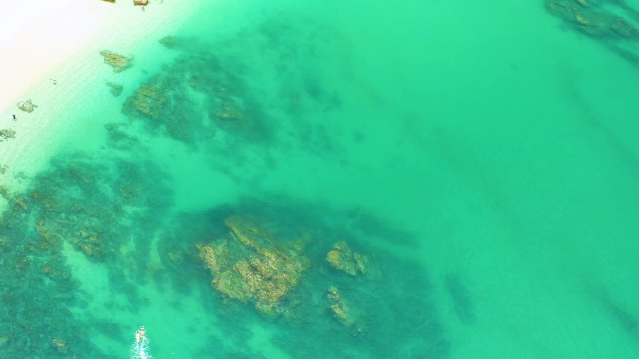 At 0:01, the Palmilla Beach frame keeps the sandy upper-left shoreline in view while large textured rocks sit beneath clear green-blue water, with no visible people.
