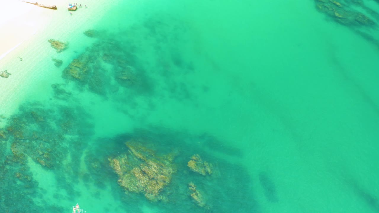 Aerial view of pale sand, turquoise water, and dark submerged rocks.