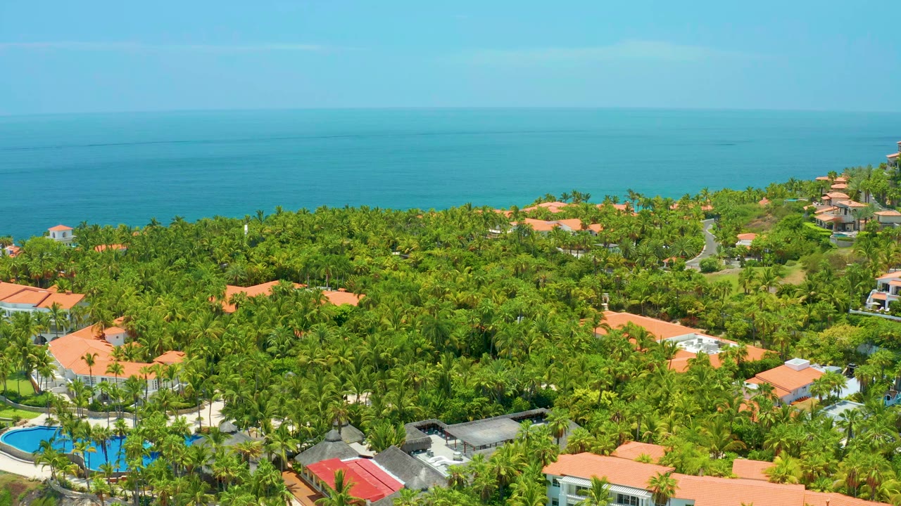 Opening aerial frame over Palmilla Beach, Baja California Sur, with dense green trees, orange roofs, a pool at lower left, blue ocean beyond, and no visible people.