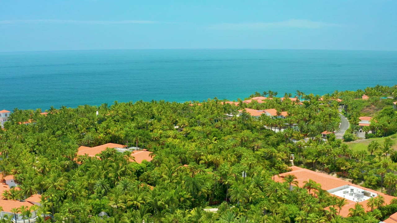 Aerial view over Palmilla Beach, Baja California Sur with dense green canopy, orange roofs near the lower edge, a blue ocean horizon, and no visible people.