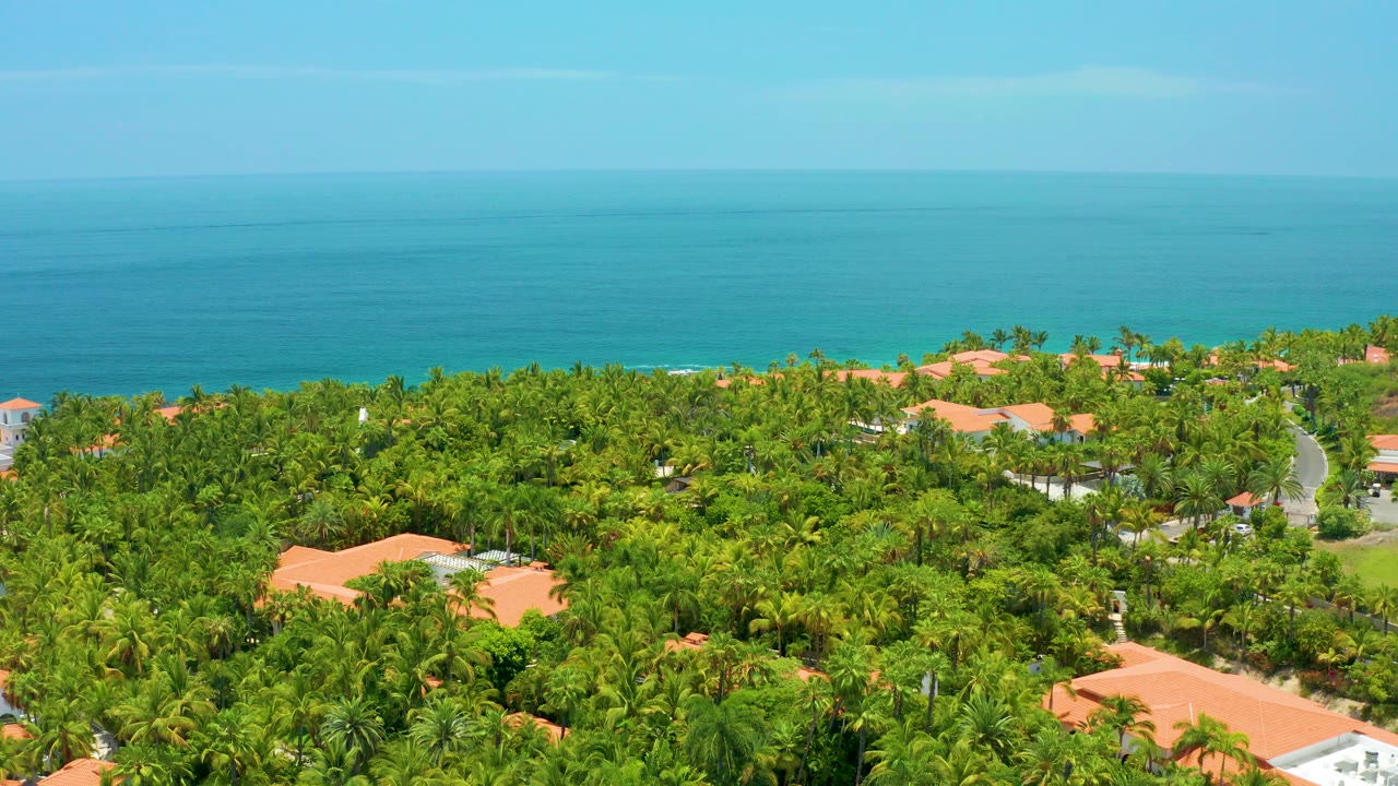 Opening aerial frame over Palmilla Beach, Baja California Sur, with dense green trees, red tile roofs, turquoise water, pale sky, and no visible people.