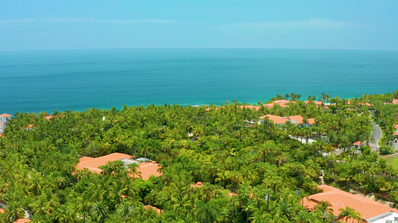 Palmilla Beach aerial view with layered green foliage, scattered red roofs, a faint shoreline, turquoise sea, and no visible people.