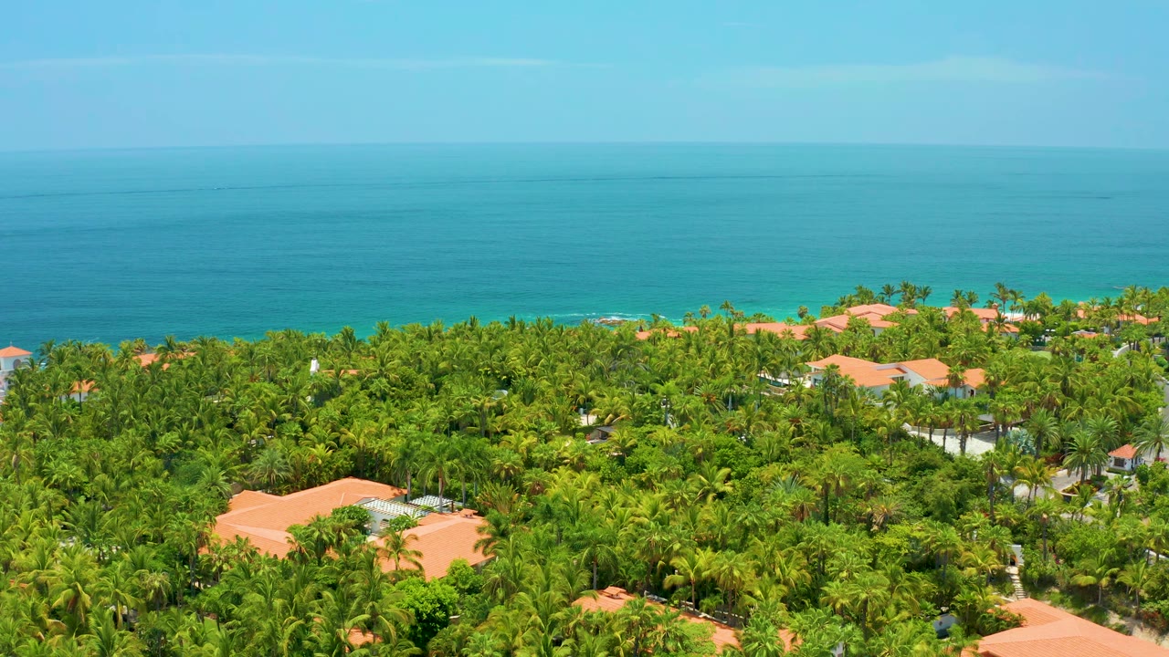 Aerial view one second into the clip showing Palmilla Beach palms and orange-roof buildings beneath a broad turquoise ocean, with no visible people.