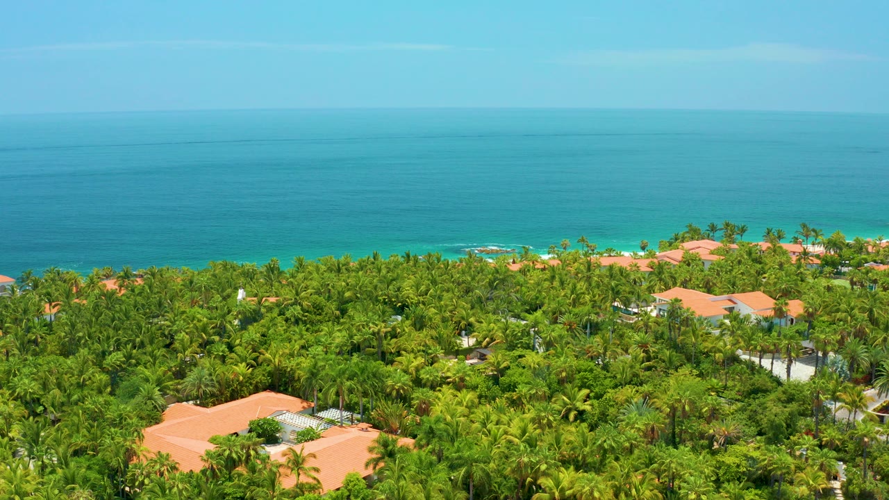 Opening aerial view over Palmilla Beach homes with orange roofs, dense green palms, blue ocean beyond, and no visible people.