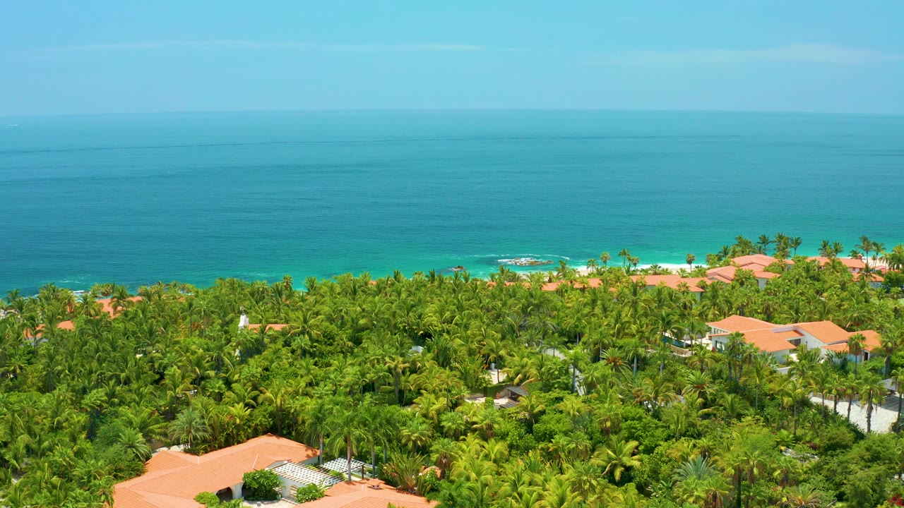 Palmilla Beach, Baja California Sur coastline seen from above with blue ocean, turquoise surf, dense palms, orange rooftops, and no visible people.