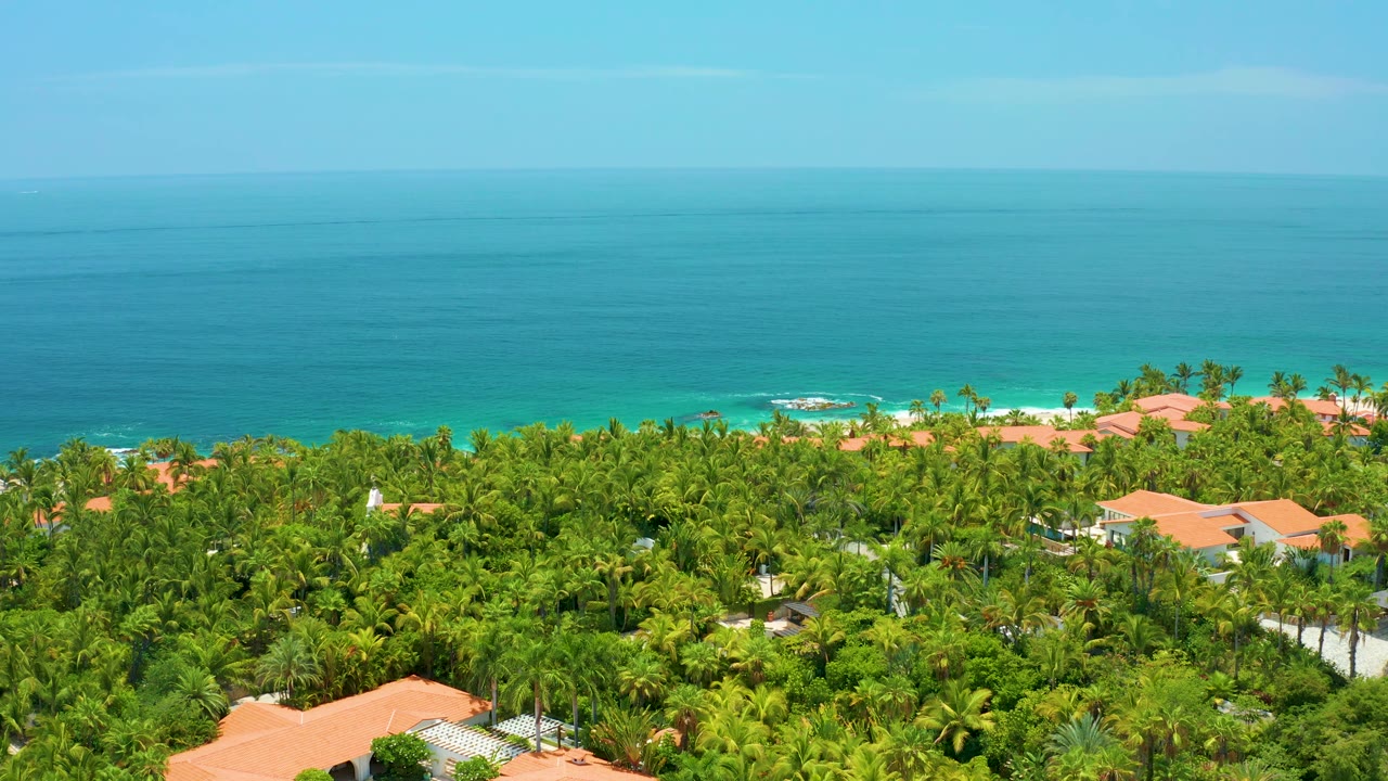 At 0:02, turquoise water and a small line of white surf appear behind dense palms and orange-roofed buildings at Palmilla Beach, with no visible people.