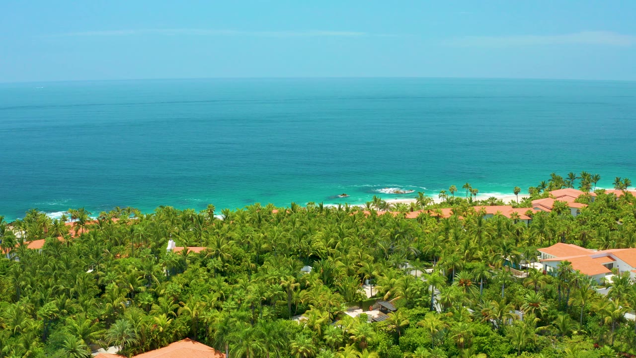 Opening aerial view of Palmilla Beach, Baja California Sur, with turquoise ocean, a strip of surf, dense palms, orange-roofed buildings, and no visible people.