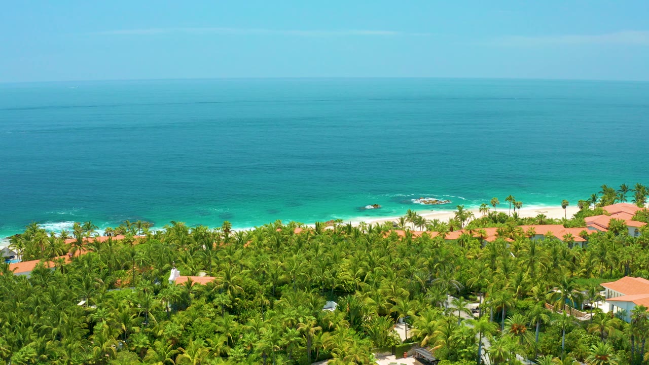 Opening aerial frame over Palmilla Beach, Baja California Sur, with dense green trees below, red tile roofs near pale sand, turquoise shore water, and no visible people.