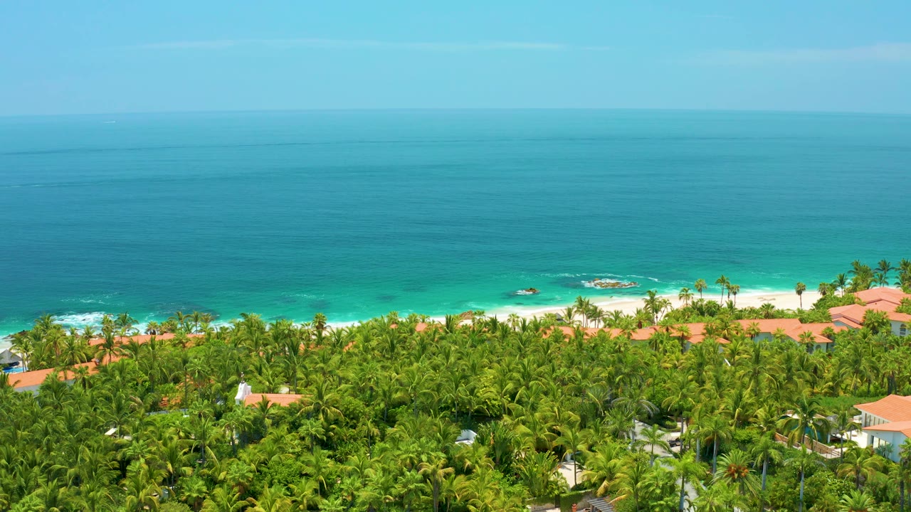 Palmilla Beach aerial view with red roofs partly visible through dense trees, pale sand along the coast, turquoise shallows, deeper blue water, and no visible people.