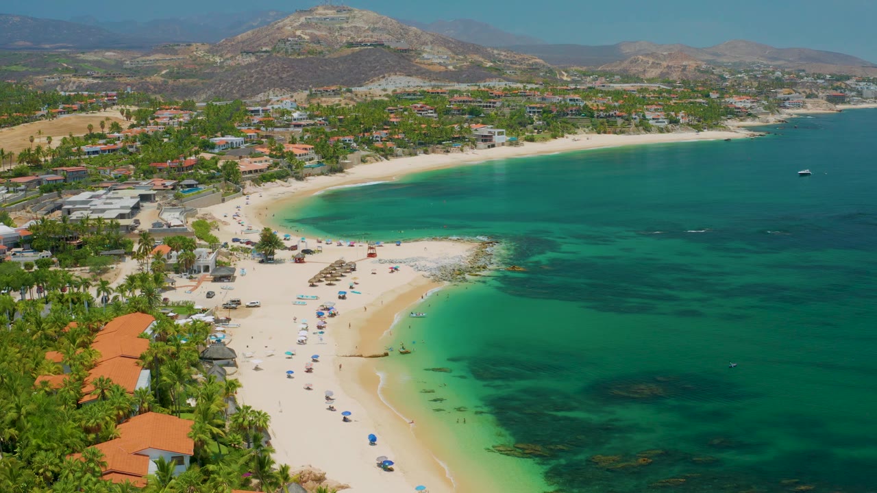Drone-like view across Palmilla Beach with a crescent of sand, umbrella rows near the waterline, palms and red roofs in the foreground, hills beyond, and no visible people.