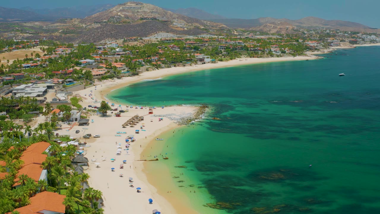 Drone view across clear green-blue water, sand beach, palms, and low hills.