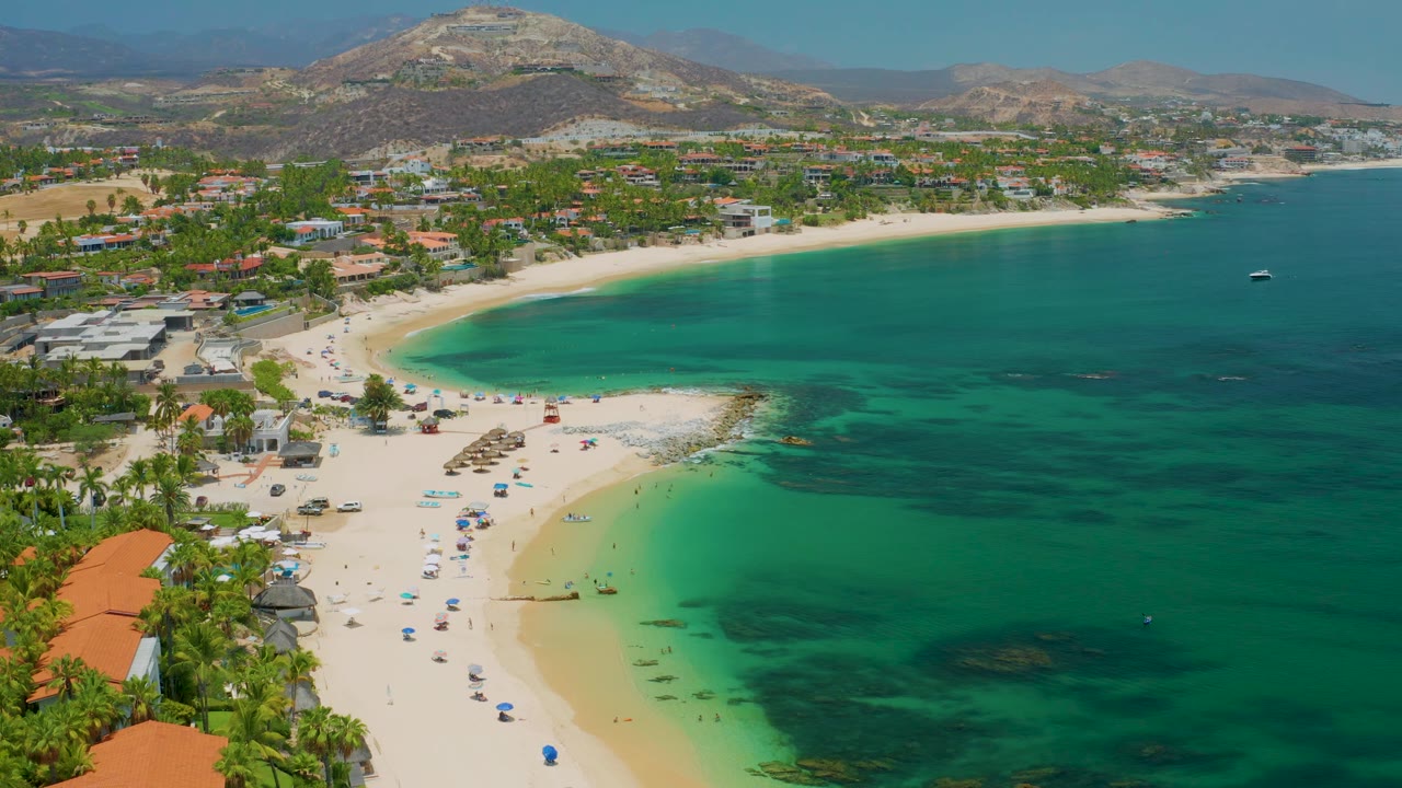 Aerial frame over Palmilla Beach with the cove centered, darker water patches offshore, rows of umbrellas on the sand, palms and rooftops along the beach, hills in the distance, and no visible people.