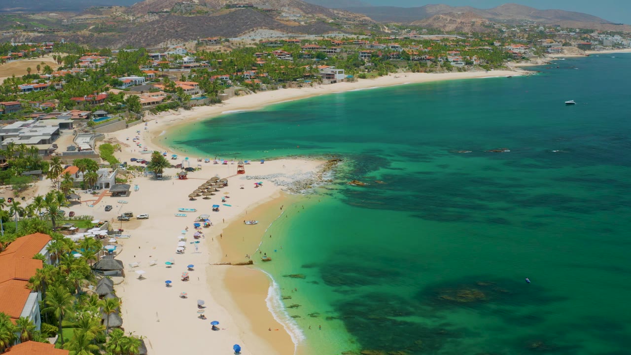 Wide aerial view of clear green-blue water beside a crescent beach with umbrellas.