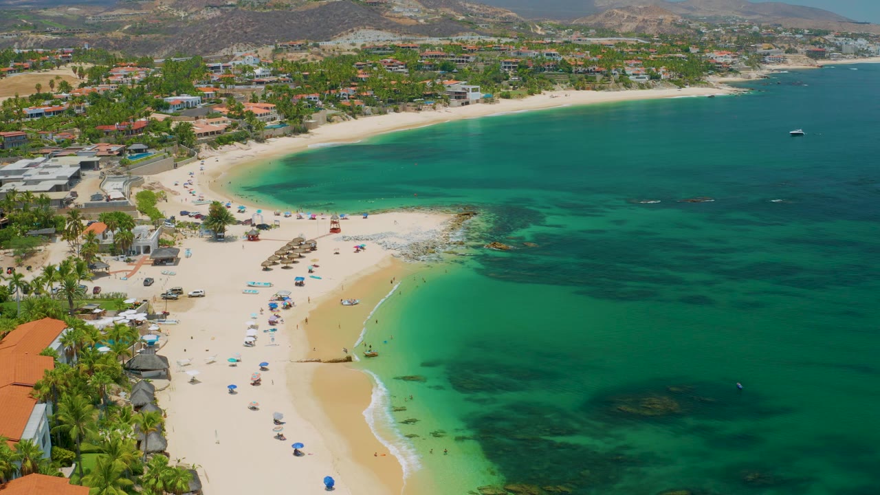 Palmilla Beach appears from above with the bay widening across the frame, umbrellas on the sand, palms and low buildings along shore, dry hills beyond, and no visible people.