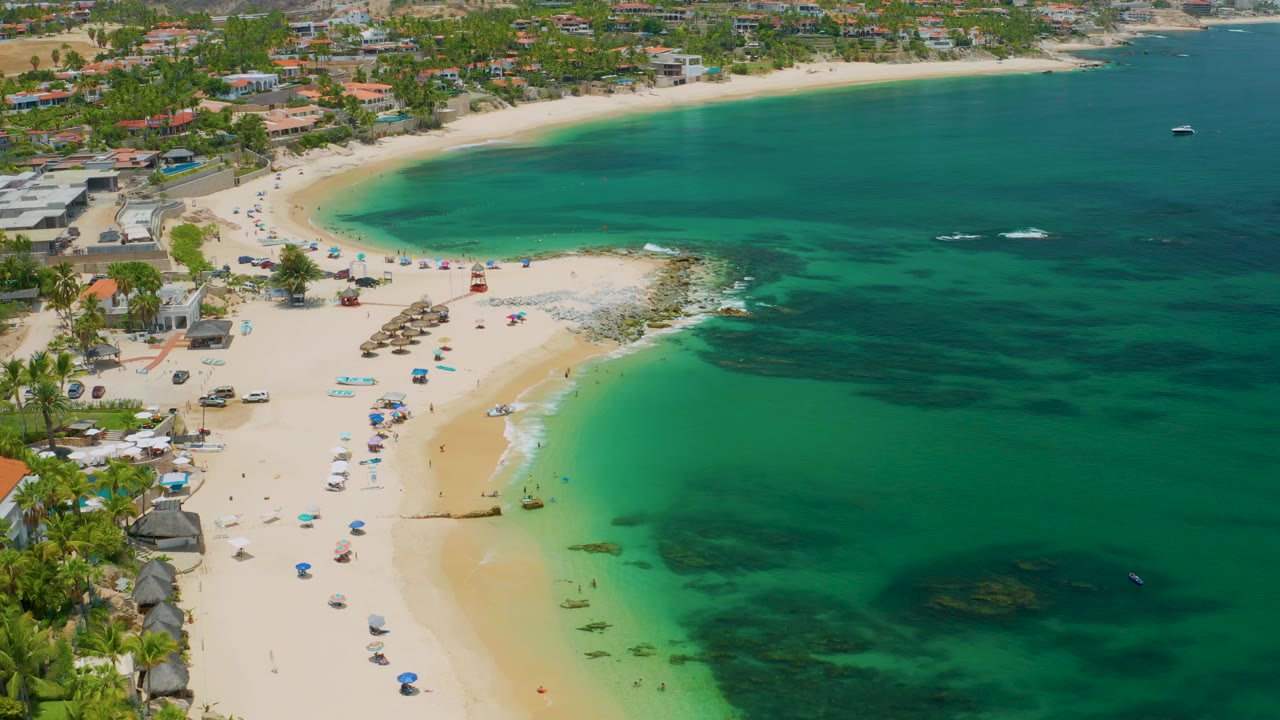 Wide aerial frame of Palmilla Beach in Baja California Sur, Mexico, with calm surf, dark underwater patches, scattered umbrellas, distant people, and coastal buildings behind the sand.