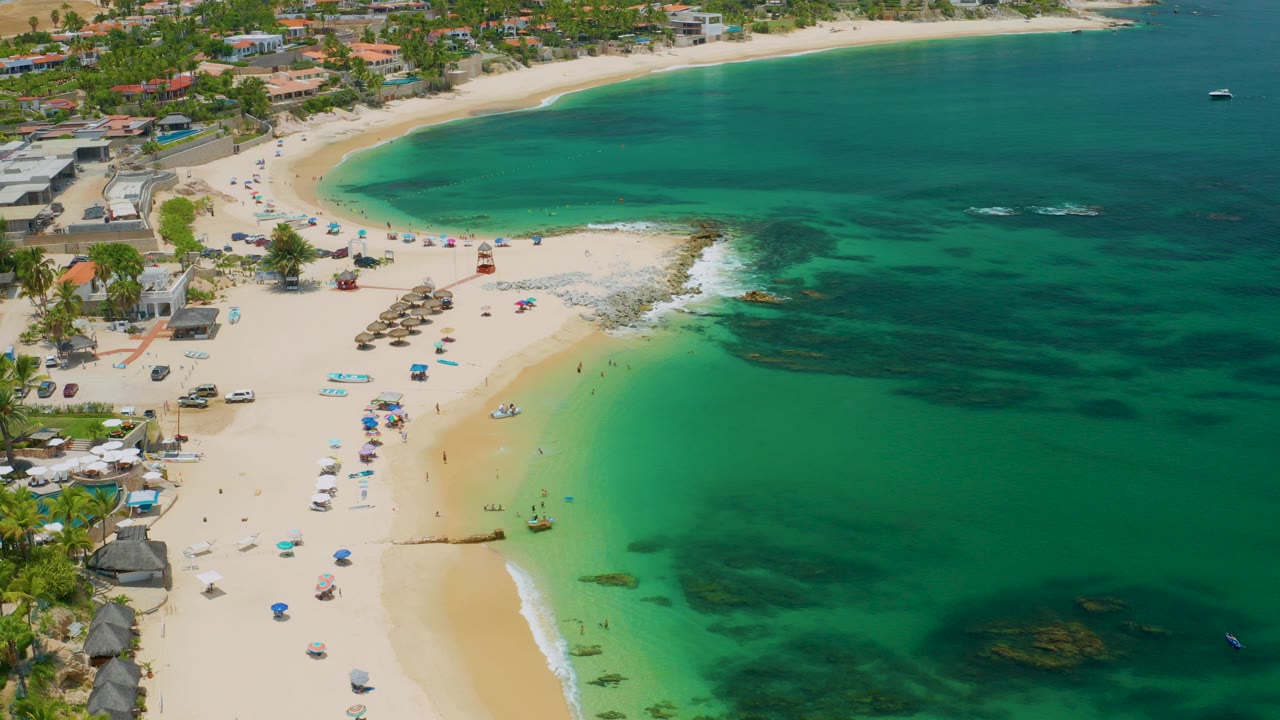 Opening aerial frame over Palmilla Beach, Baja California Sur, with pale sand, umbrellas, small beachgoers, beachfront buildings, and green-blue water curving around the shore.