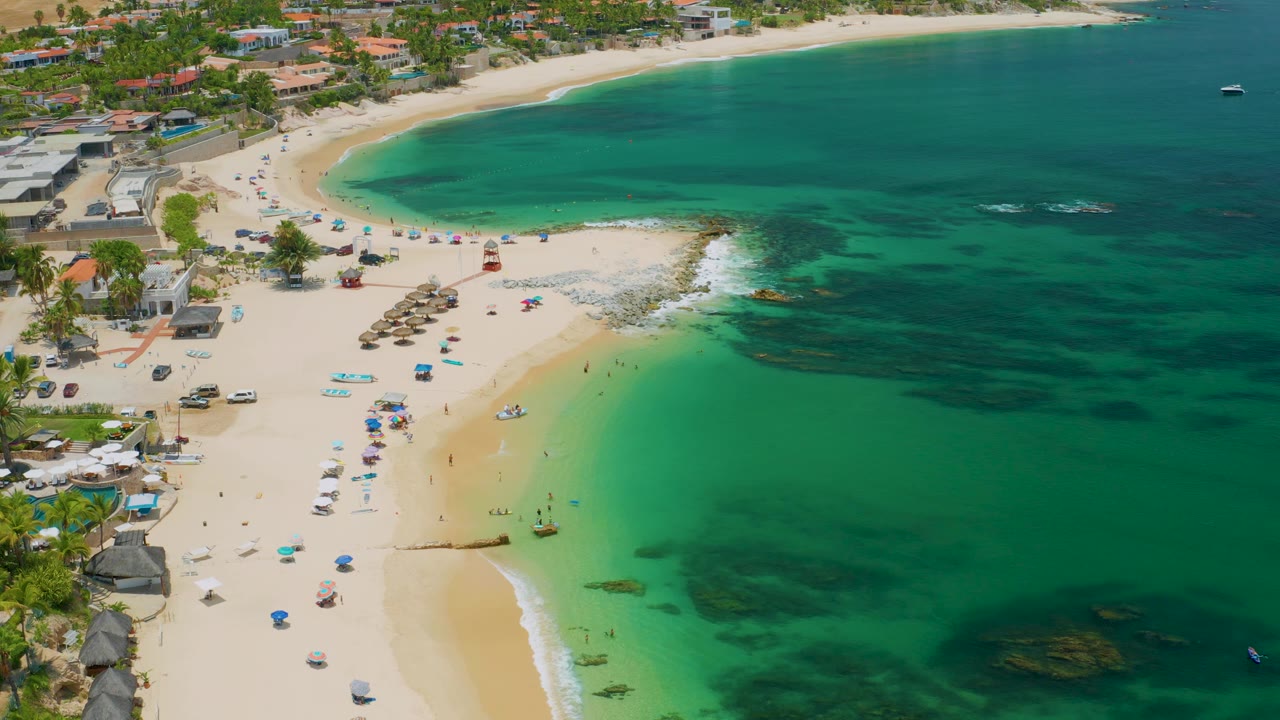 Palmilla Beach aerial frame with more open water to the right, a sandy swimming area below, tiny people near the waterline, and beachfront buildings set back from the sand.