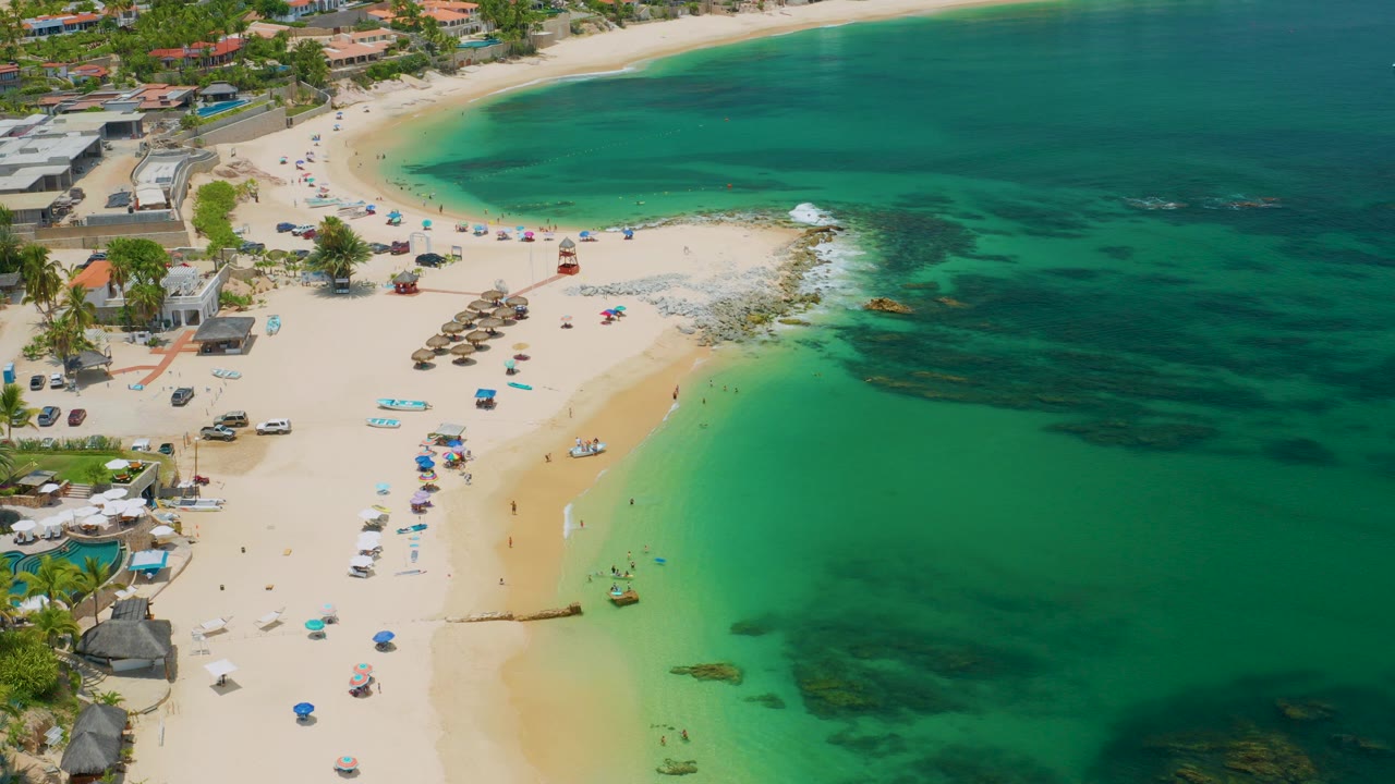 Aerial view over Palmilla Beach in Baja California Sur, showing pale sand, beach umbrellas, shoreline buildings, and green-blue water with tiny distant beachgoers.