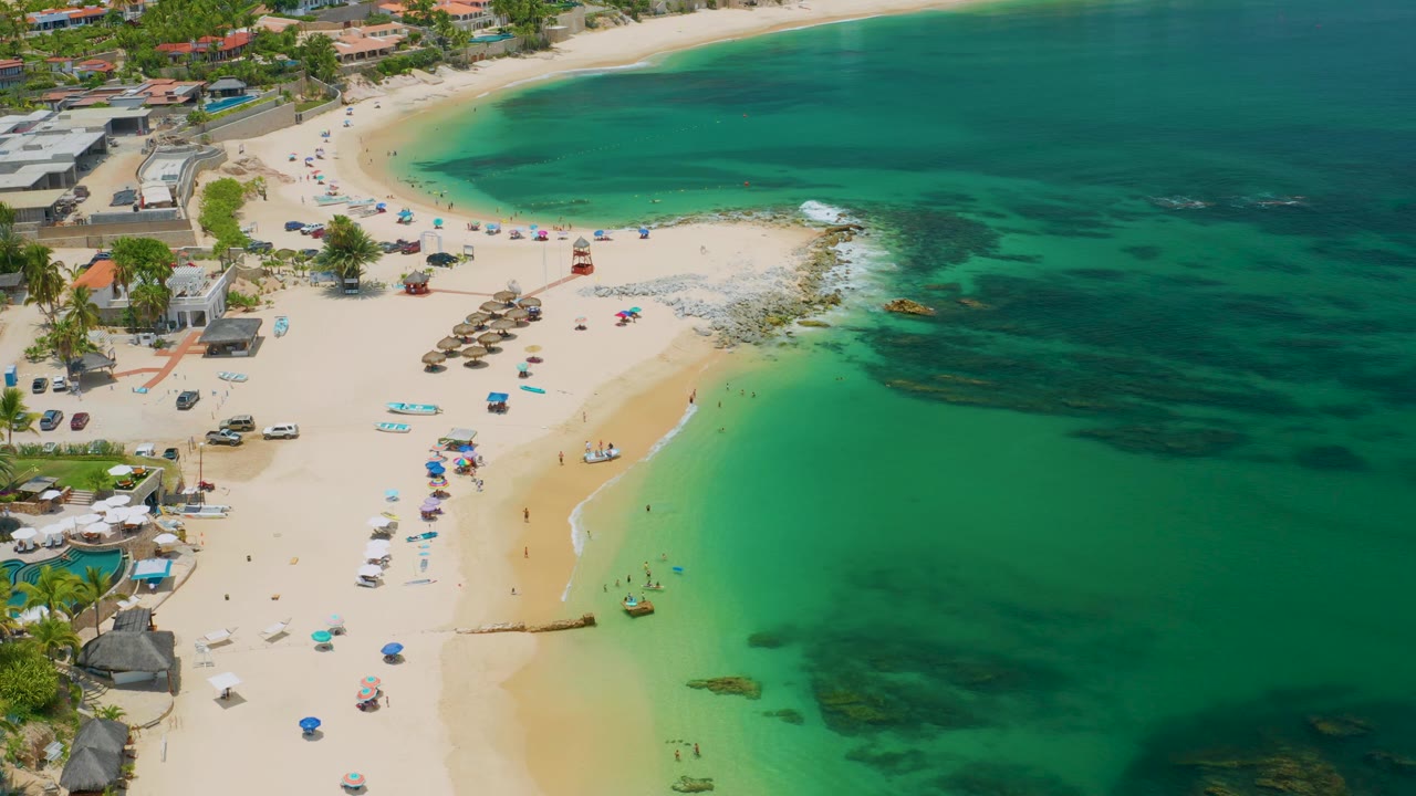 Palmilla Beach from above, with a broad sweep of pale sand at left, clear green water at right, and a line of umbrellas and small beach items near the shore.