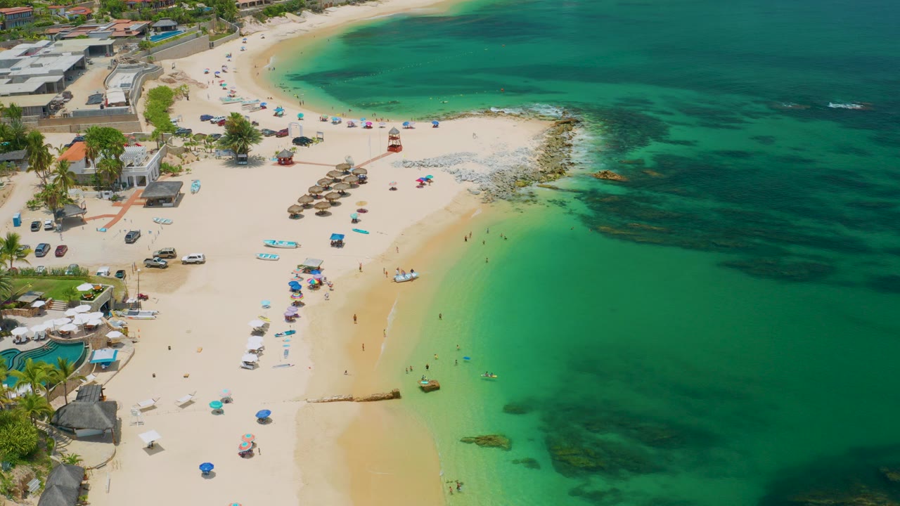 Opening aerial frame over Palmilla Beach in Baja California Sur, with resort buildings at left, pale sand through the center, clear green water at right, beach umbrellas, shade structures, and no identifiable people.