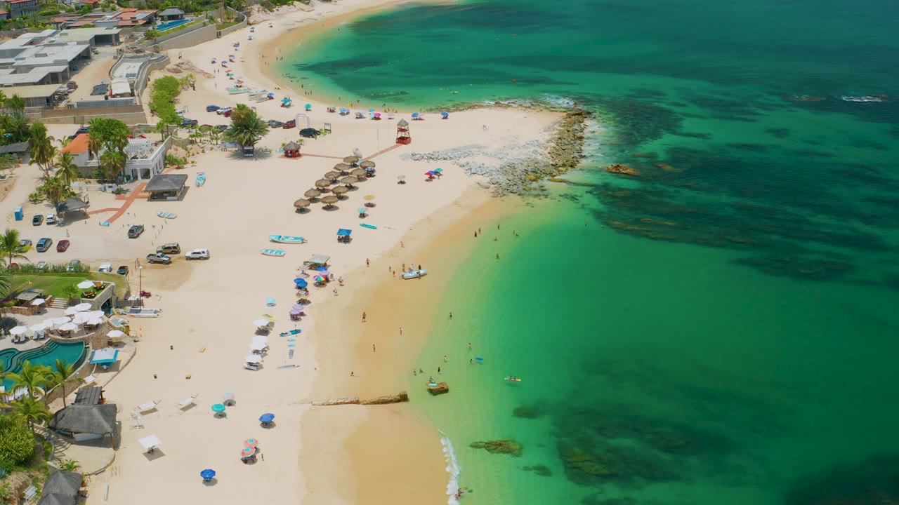 Aerial shoreline with beach umbrellas, palapa shades, resort buildings, and clear water.