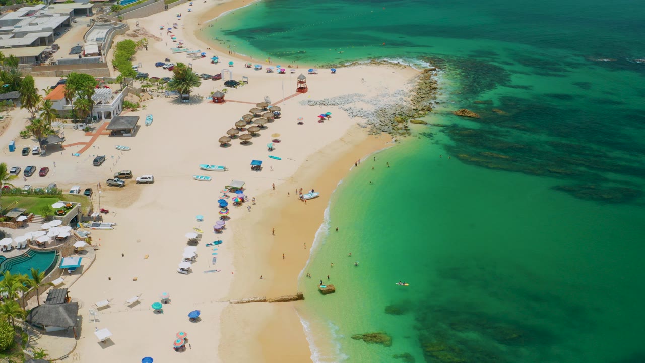 Aerial frame over Palmilla Beach in Baja California Sur, showing pale sand, turquoise shallows, darker water patches, umbrellas, small boats, and tiny beachgoers visible from above.