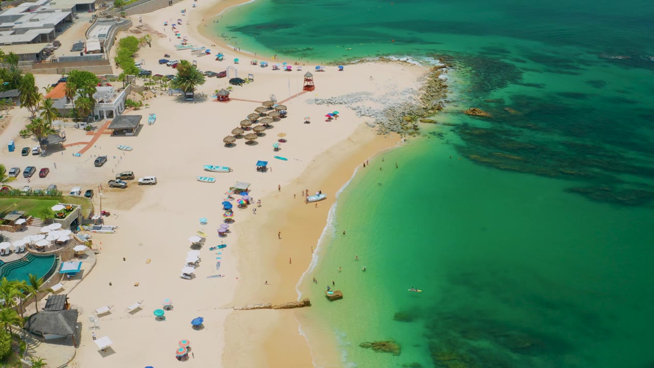 Palmilla Beach appears from above with resort-edge structures at the upper left, rows of umbrellas and chairs on the sand, a small boat near the waterline, and clear shallow water with darker patches.