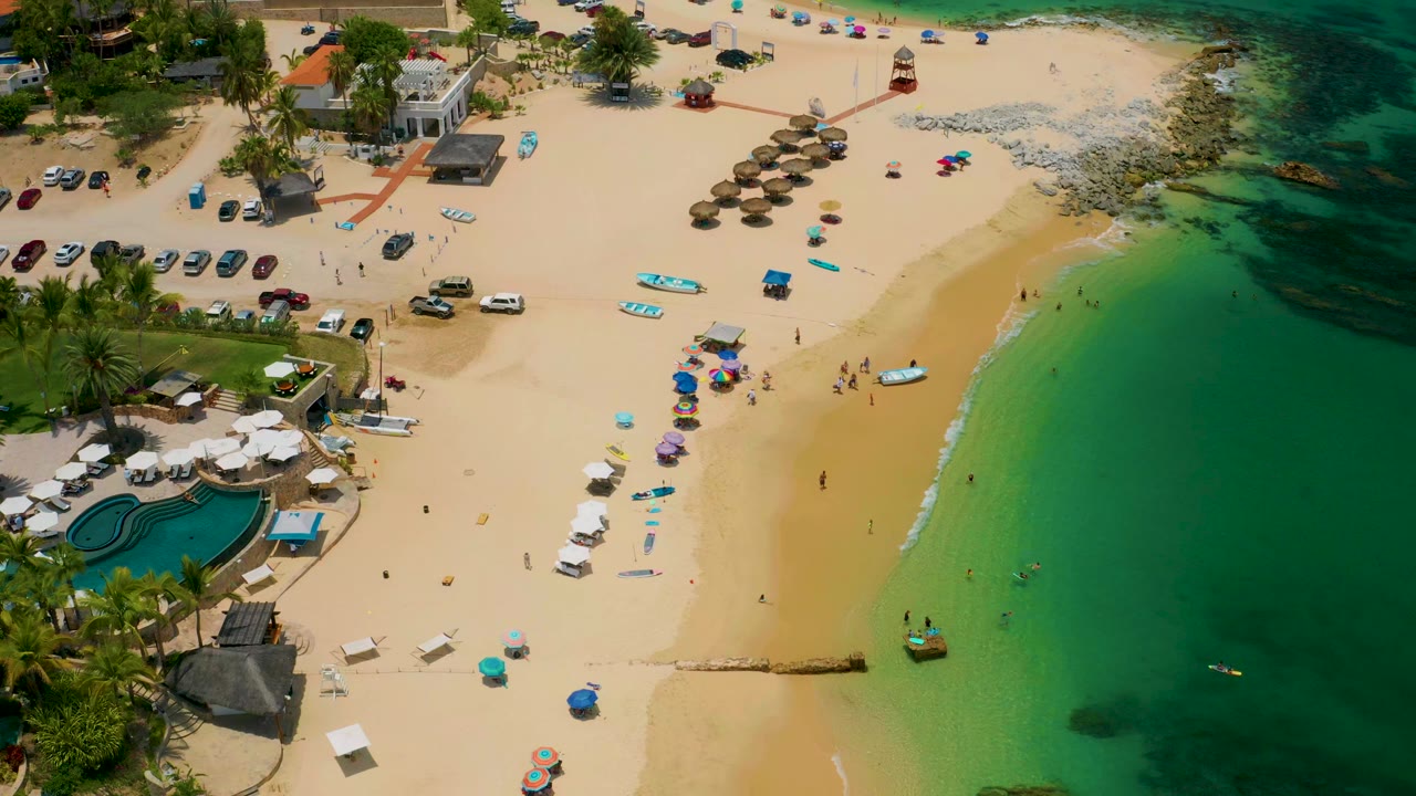 High aerial view of Palmilla Beach in Baja California Sur with parked cars and palms at left, umbrellas on the sand, small people near the shoreline, and turquoise water filling the right side.