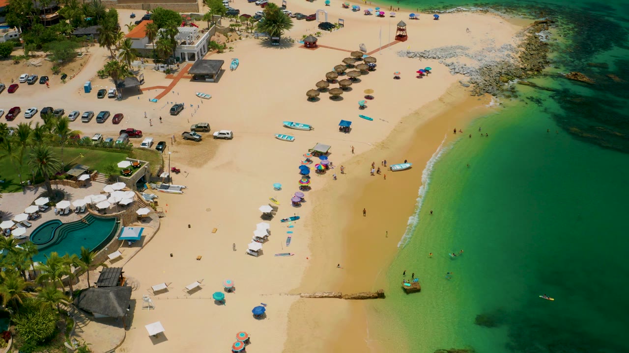 High oblique view of Palmilla Beach with palapa-style umbrellas near the upper center, scattered beachgoers below, parked cars beside trees, and darker rocky water at the upper right.