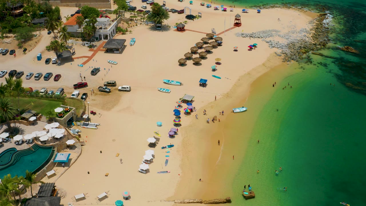 Aerial view of Palmilla Beach with a broad strip of sand, people moving in small groups, umbrellas and loungers near the lower center, thatched shade structures near the upper beach, and clear green water along the right.