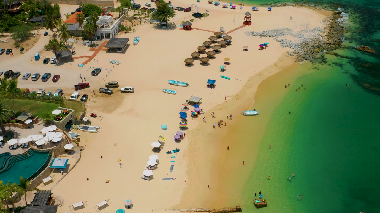 Aerial beach view with umbrellas, swimmers, parked cars, palms, and a pool area.