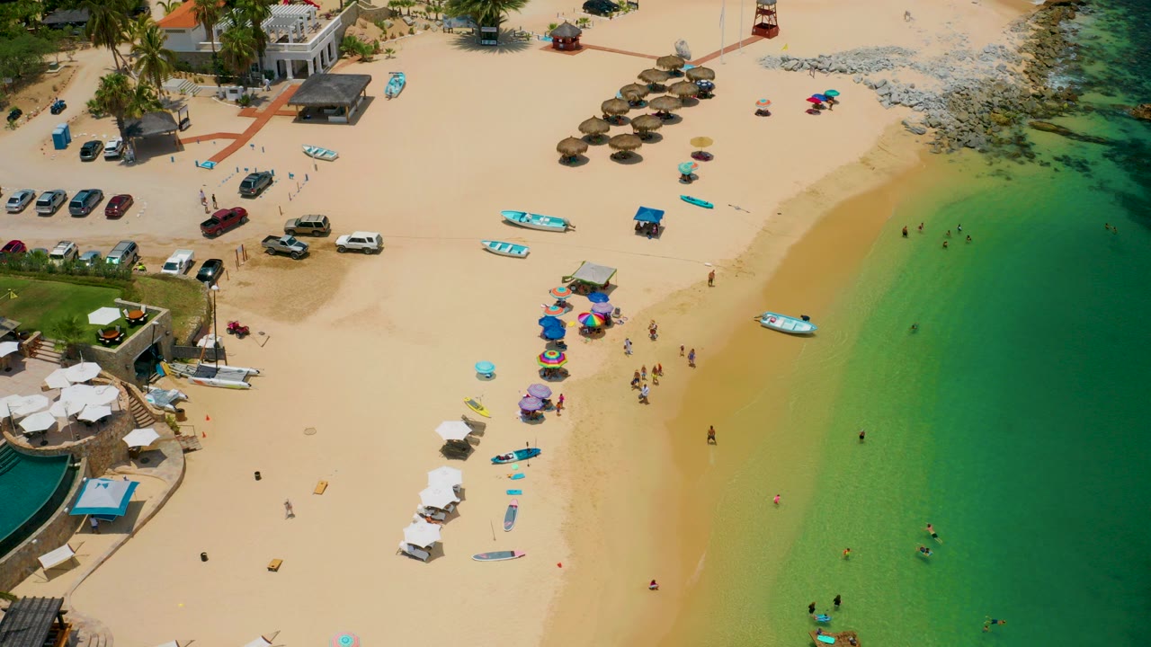 High aerial view of Palmilla Beach, Baja California Sur, with pale sand, colored umbrellas, small boats, parked cars, beachfront buildings, and swimmers in green water.