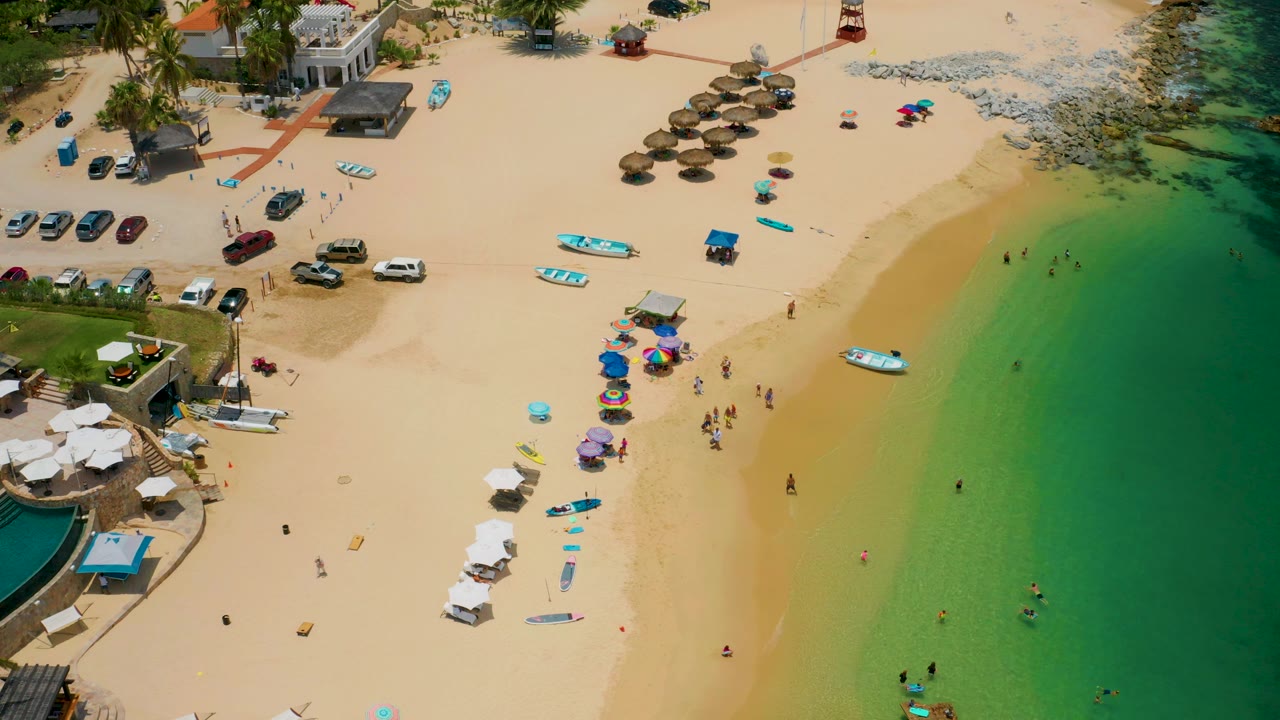 Overhead shoreline view with beach umbrellas, swimmers, boats, and parked vehicles.