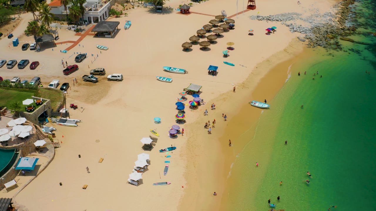 Palmilla Beach from above, with the green water occupying the right side, a small boat close to the shoreline, colored umbrellas on the sand, and beachgoers visible as small figures.